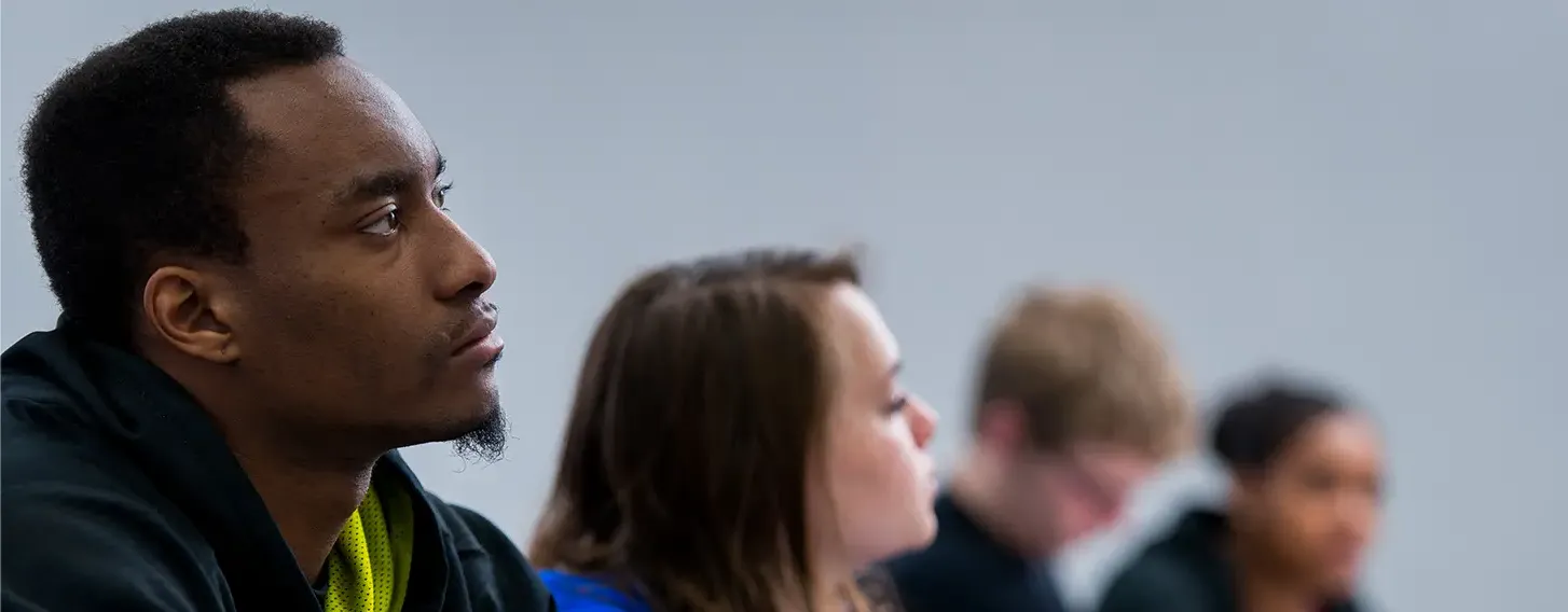 A diverse group of students sitting at desks in a classroom, listening to the teacher and taking notes.